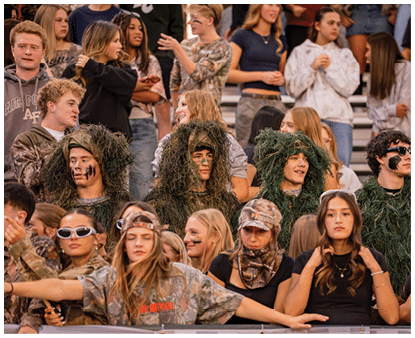 A group of people in camouflage and face paint are sitting and standing closely together in bleachers, some wearing ghillie suits and others in casual clothing, appearing to be at an outdoor event. Description generated by AI