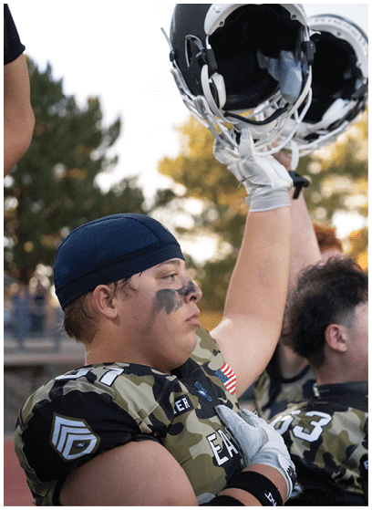 People wearing camouflage football uniforms raise their helmets in the air during an outdoor event. Description generated by AI