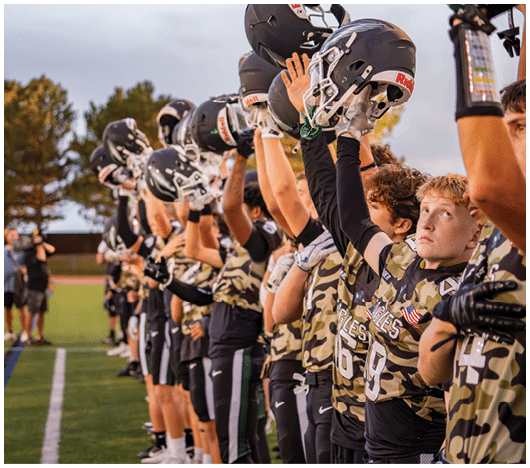 A line of people wearing camouflage sports uniforms stand on a field holding up black football helmets in a gesture of respect or celebration. Description generated by AI