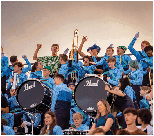 A group of people in blue uniforms are playing musical instruments and cheering in a stadium setting while others in casual clothing watch from the stands. Description generated by AI