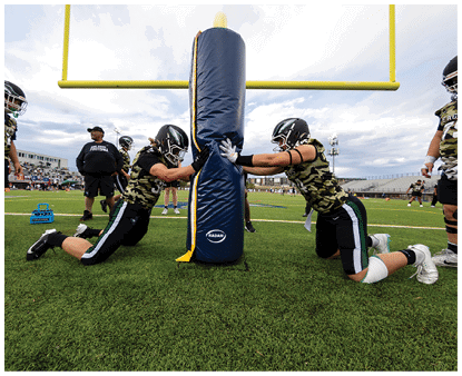 Two people in camouflage sports uniforms and helmets push against a padded tackling dummy on a football field under a goalpost. Description generated by AI