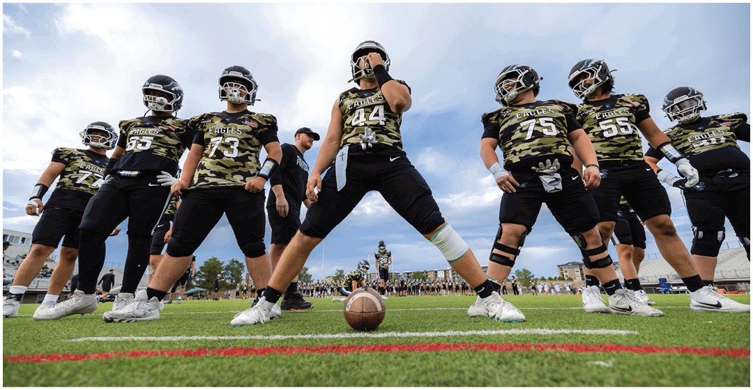 A group of adults in camouflage sports jerseys and helmets stand on a football field with one person in the center positioned behind a football on the ground. Description generated by AI