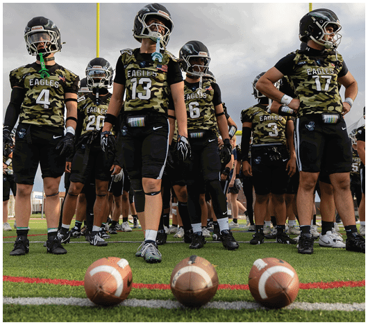 A group of people in camouflage sports uniforms and helmets stand on a football field behind three footballs lined up on the grass. Description generated by AI