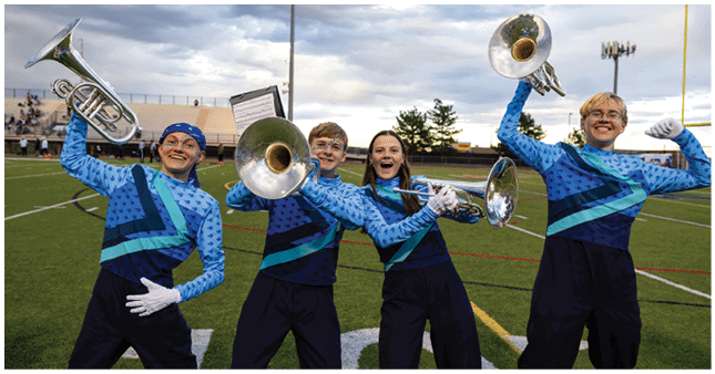 Four people wearing matching blue and black band uniforms enthusiastically hold brass instruments on a sports field during what appears to be a band performance or practice. Description generated by AI