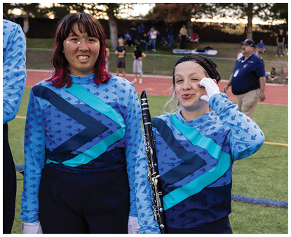 Two people wearing matching blue and teal band uniforms stand on a field, with one holding a clarinet and the other smiling. Description generated by AI