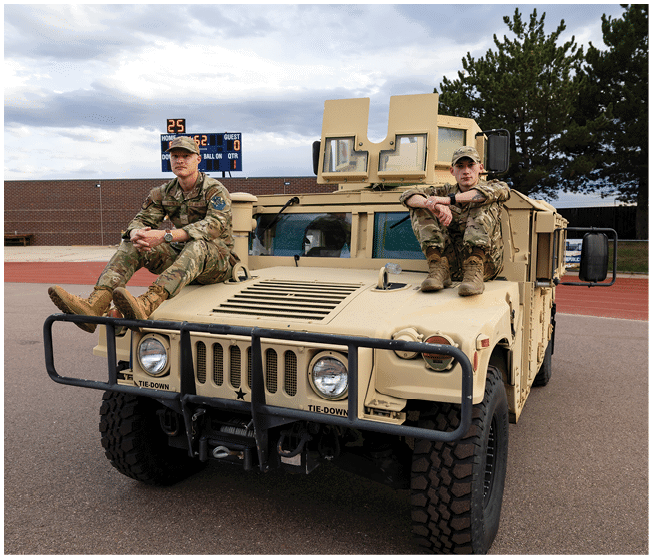 Two people in military uniforms sit on the hood of a tan armored vehicle parked on a paved area near a scoreboard and trees. Description generated by AI