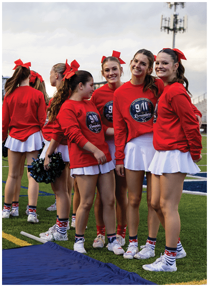A group of people wearing red long-sleeve shirts with 9/11 remembrance logos, white skirts, red bows in their hair, and patriotic socks are standing on a sports field, some smiling and holding pom-poms. Description generated by AI