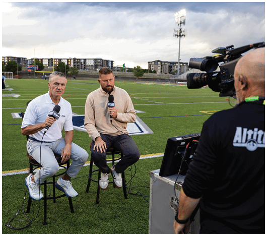 Two adults sit on stools on a sports field holding microphones while being recorded by a camera operator. Description generated by AI