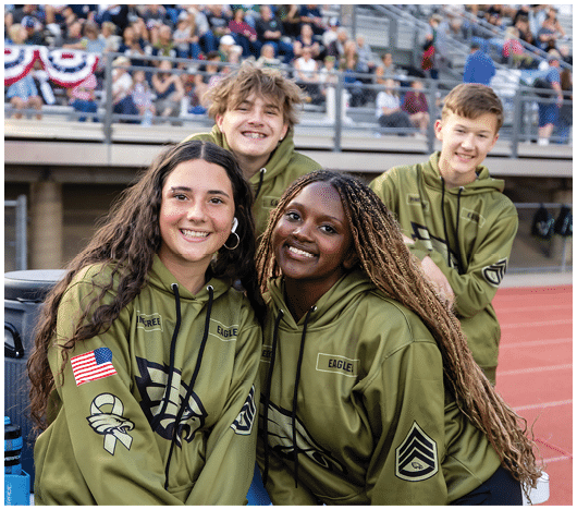 Four smiling people wearing matching green hoodies with patches are posing together at an outdoor sports venue with a crowd in the background. Description generated by AI