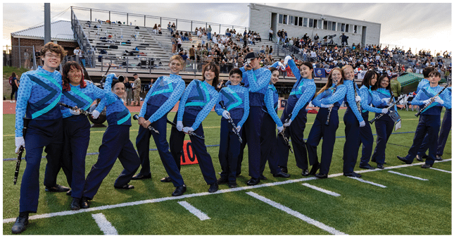 A group of people in matching blue and navy uniforms stand in a line on a football field holding musical instruments with a crowd seated in bleachers behind them. Description generated by AI