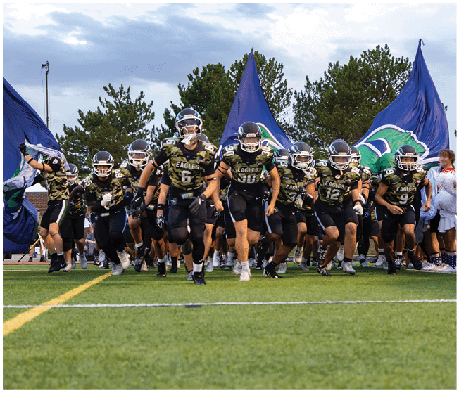 A group of football players in black uniforms with helmets are running onto a field through large blue flags held by people on either side. Description generated by AI