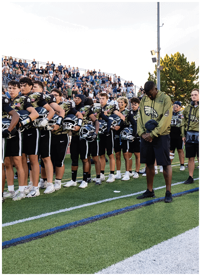 A group of people wearing matching green jackets and black shorts stand in a line on a sports field near a sideline marker and footballs. Description generated by AI