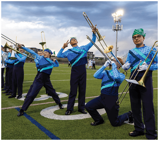 People in matching blue and green band uniforms pose with trombones on a football field under a cloudy sky. Description generated by AI