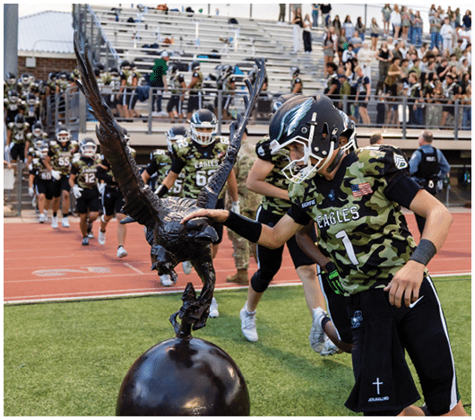 A person in a football uniform with a helmet is touching a large eagle statue on a sports field while other players and spectators are visible in the background. Description generated by AI