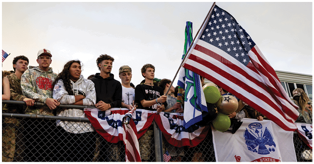 A group of people stand behind a fence decorated with patriotic bunting while one person holds an American flag and balloons. Description generated by AI