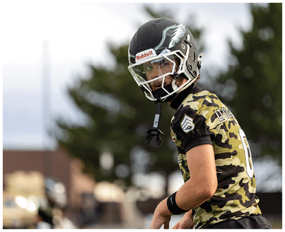 A person wearing a camouflage sports jersey and a football helmet is looking back while standing outdoors near trees and buildings. Description generated by AI