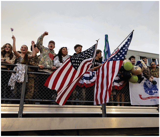People standing behind a fence enthusiastically waving American flags and holding balloons during an outdoor event. Description generated by AI