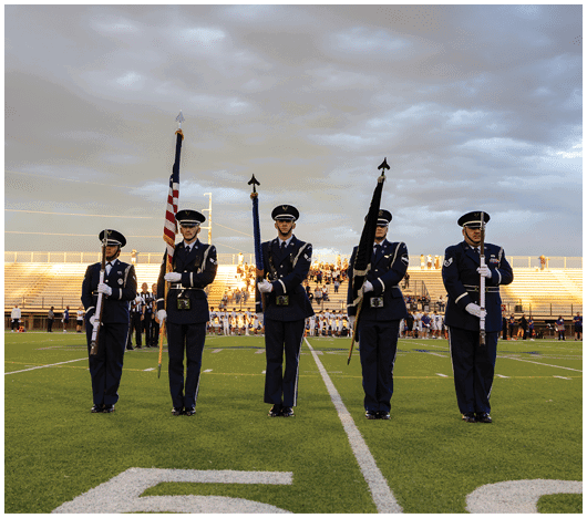 Five people in formal military uniforms stand in a line on a football field holding flags and rifles with a crowd and bleachers in the background under a cloudy sky. Description generated by AI