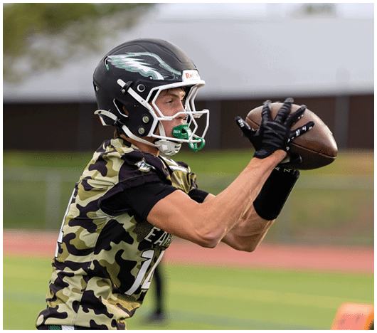 A person wearing a black football helmet with a green eagle logo and a camouflage jersey is catching a football on a field. Description generated by AI