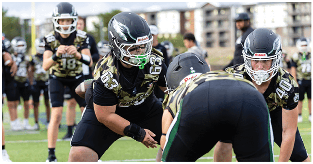 People wearing football helmets and camouflage jerseys are positioned on a field preparing for a play during a game. Description generated by AI