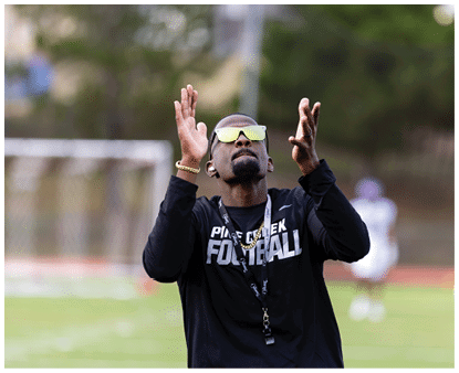 A person wearing sunglasses and a black long-sleeve shirt with “FOOTBALL" written on it is raising their hands outdoors on a sports field. Description generated by AI