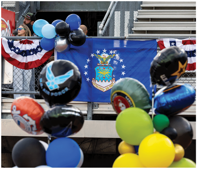 A decorated outdoor event area features a blue flag with an emblem, patriotic bunting, and clusters of colorful balloons. Description generated by AI