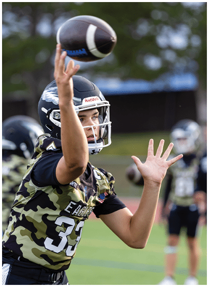 A person wearing a camouflage sports jersey and helmet is throwing a football on a grassy field with other players in the background. Description generated by AI