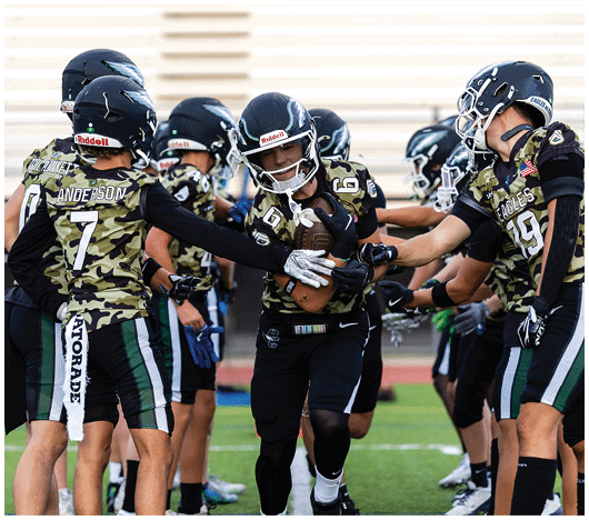 A group of people wearing camouflage football uniforms and helmets are gathered on a field with one person holding a football while others reach out to them. Description generated by AI