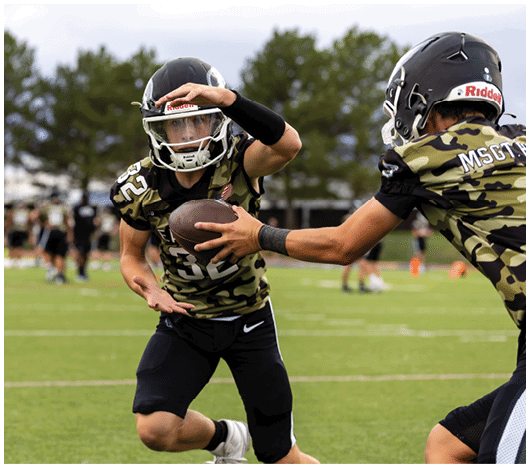 Two people wearing camouflage football uniforms and helmets are on a field with one handing a football to the other. Description generated by AI
