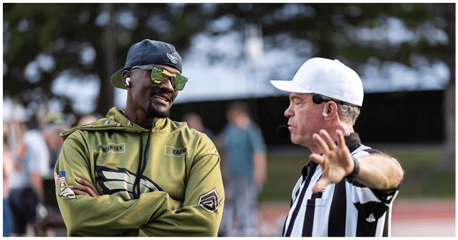 Two adults are engaged in conversation outdoors, one wearing a green hoodie and sunglasses with earbuds, and the other in a black-and-white striped referee shirt gesturing with a raised hand. Description generated by AI