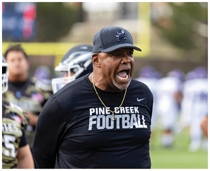 A person wearing a Pine Creek Football shirt and cap is shouting passionately on a sports field with blurred players in the background. Description generated by AI