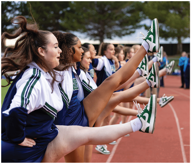 A line of adults in cheerleading uniforms are performing a high kick on a running track outdoors. Description generated by AI