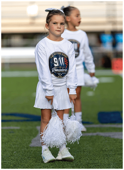 A child wearing a white cheerleading outfit with a 9/11 remembrance logo holds white pom-poms while standing on a sports field with another child in the background. Description generated by AI