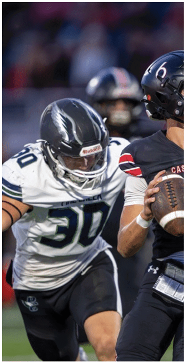 A person wearing a black football uniform with the number six is holding a football and preparing to throw it while two opposing players in white uniforms attempt to block. Description generated by AI