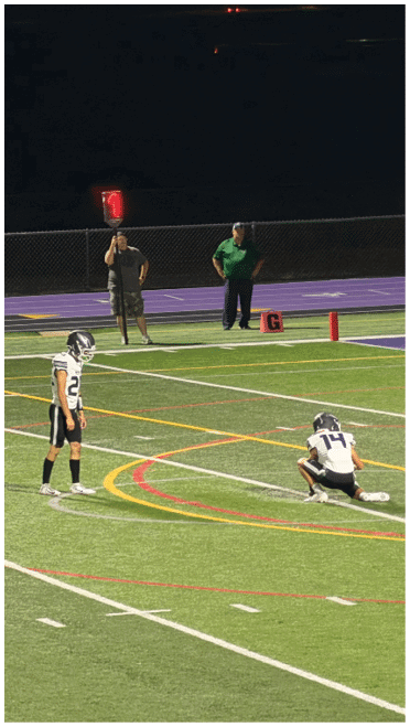 Two people in football uniforms prepare for a play on a well-lit field while two adults stand near the sideline at night. Description generated by AI