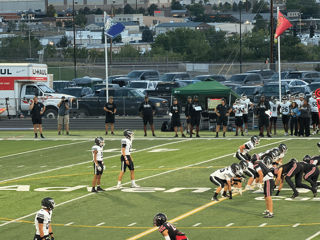 A group of people in football uniforms are positioned on a football field preparing for a play while several people stand on the sidelines near a green tent and parked vehicles in the background. Description generated by AI
