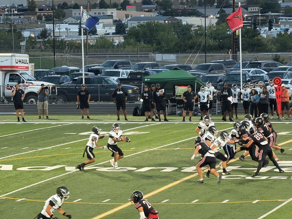 People in football uniforms are actively playing a game on a green field with a crowd and parked cars in the background. Description generated by AI