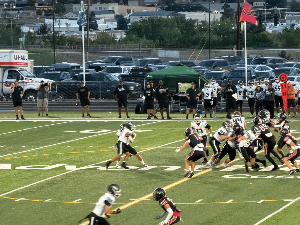 People in football uniforms are actively playing a game on a field with coaches and other players standing on the sidelines near a chain marker. Description generated by AI