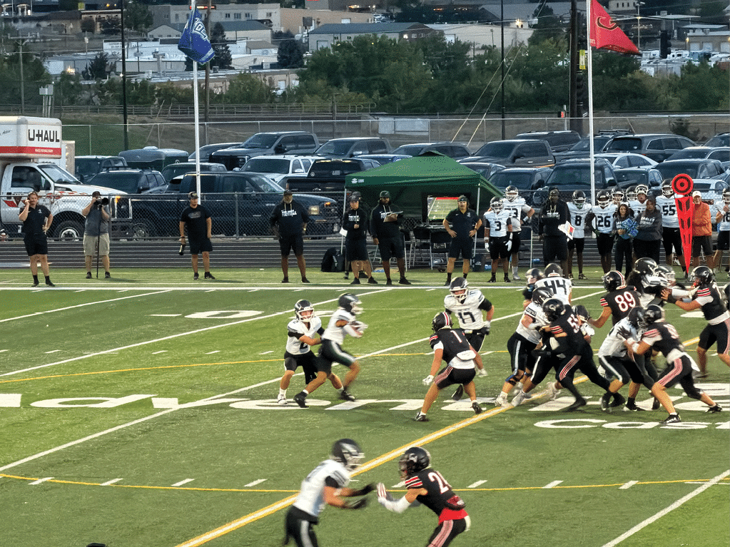 A group of people in football uniforms are actively playing a game on a field with spectators and team members standing on the sidelines. Description generated by AI