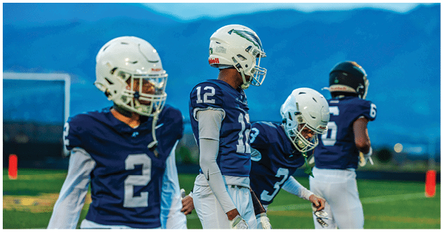 Four people wearing blue football uniforms and helmets stand on a field with mountains in the background. Description generated by AI