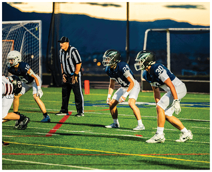 Three people in football uniforms and helmets are positioned on a field with a referee standing behind them during a game at dusk. Description generated by AI