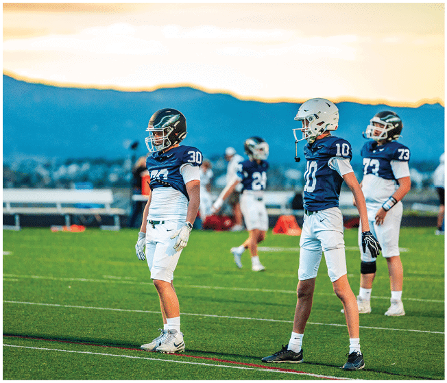 Three people in football uniforms stand on a green field with mountains and a sunset sky in the background. Description generated by AI
