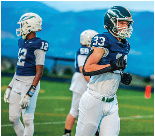 Three people wearing football uniforms and helmets stand on a field with mountains in the background during what appears to be a practice or warm-up session. Description generated by AI