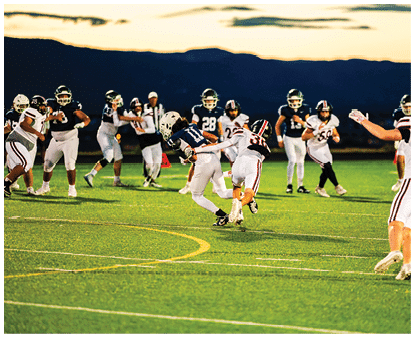 People wearing football uniforms are actively playing on a well-lit field during a game at night. Description generated by AI