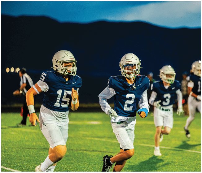 Two people wearing football uniforms and helmets are running on a field at night with a referee in the background. Description generated by AI