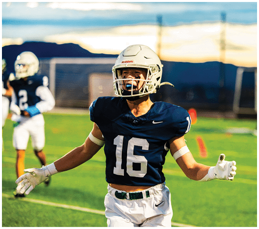 A person wearing a football helmet and dark jersey with the number 16 is celebrating on a football field with another player in the background. Description generated by AI