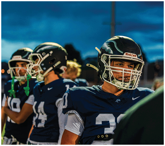 People wearing football helmets and uniforms stand together on a field during a game or practice at dusk. Description generated by AI