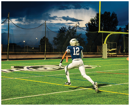 A person wearing a blue football jersey with the number 12 is running on a green football field near the goalpost during dusk. Description generated by AI