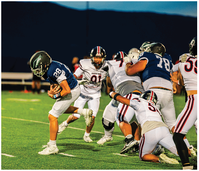 A person wearing a blue football uniform is running with the ball while several people in white football uniforms are attempting to catch them on a lit outdoor field at dusk. Description generated by AI