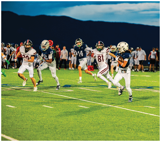 Several people in football uniforms are actively running on a well-lit field during a game at dusk with a dark mountain silhouette in the background. Description generated by AI